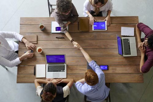 Overhead view of a team around a wooden table with laptops and tablets, two people shaking hands