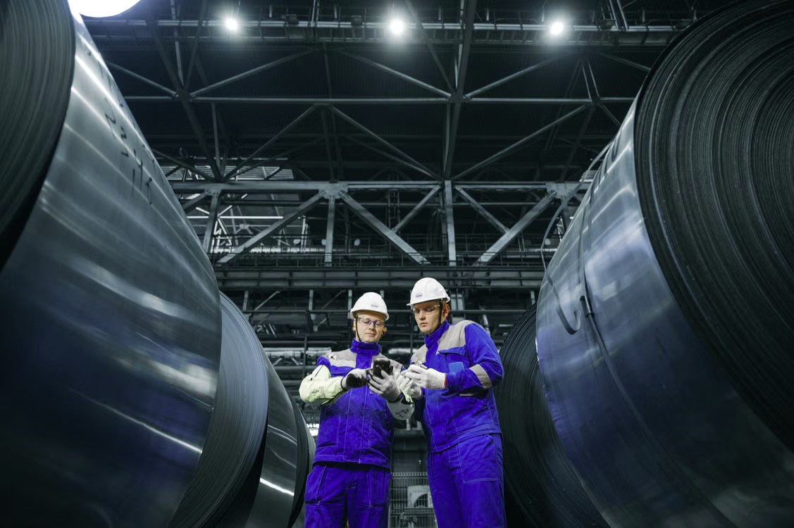 Two industrial workers in hard hats reviewing inventory between large rolls of steel coil